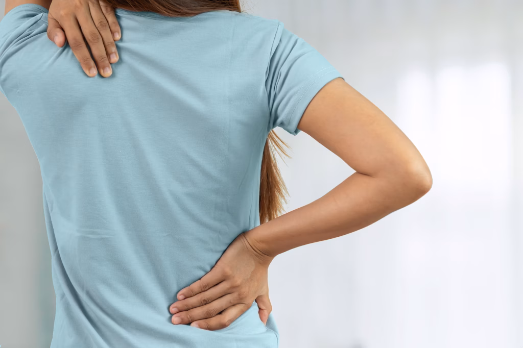Spinalkanalstenose: Rückansicht einer jungen Frau im hellblauen T-Shirt, die sich mit der rechten Hand in die Taille greift und mit der linken Hand das Schulterblatt berührt.